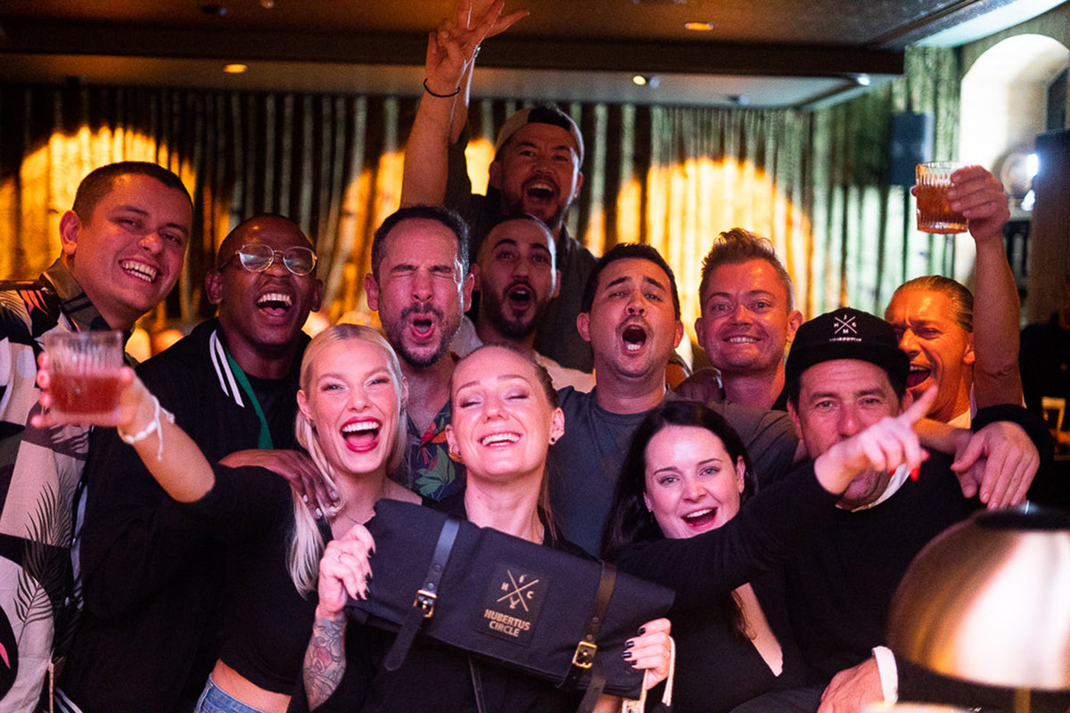A group of ten friends celebrating together in a dimly lit bar or club, holding drinks and smiling, with one person holding a black designer handbag featuring a gold logo.