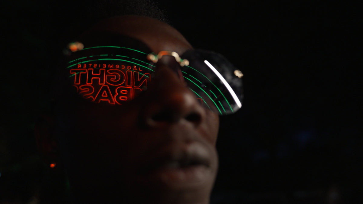 A close-up shows the face of a person whose sunglasses reflect the glowing reflection of green and red neon light with lettering in the darkness.