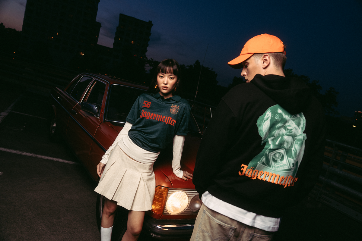 Two models posing in front of a vintage red car at dusk. One model wears a black Jägermeister Waldi hoodie, the other one wears a cream pleated skirt, and a green Jägermeister jersey.