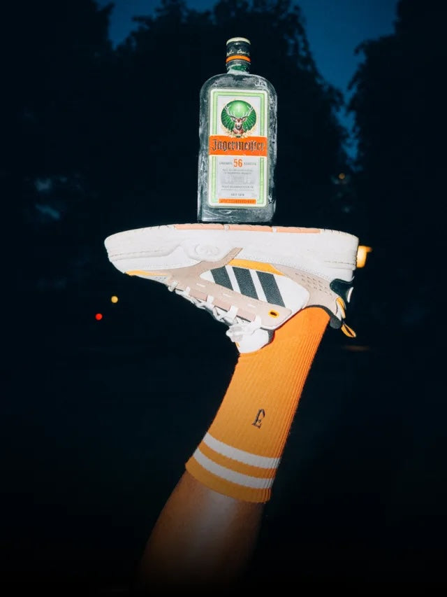 A white and orange sneaker rests on a bright orange ribbed sock, which is balanced on the edge of a hand holding a bottle of Jägermeister. The background is a very dark, nighttime outdoor setting with silhouetted trees.