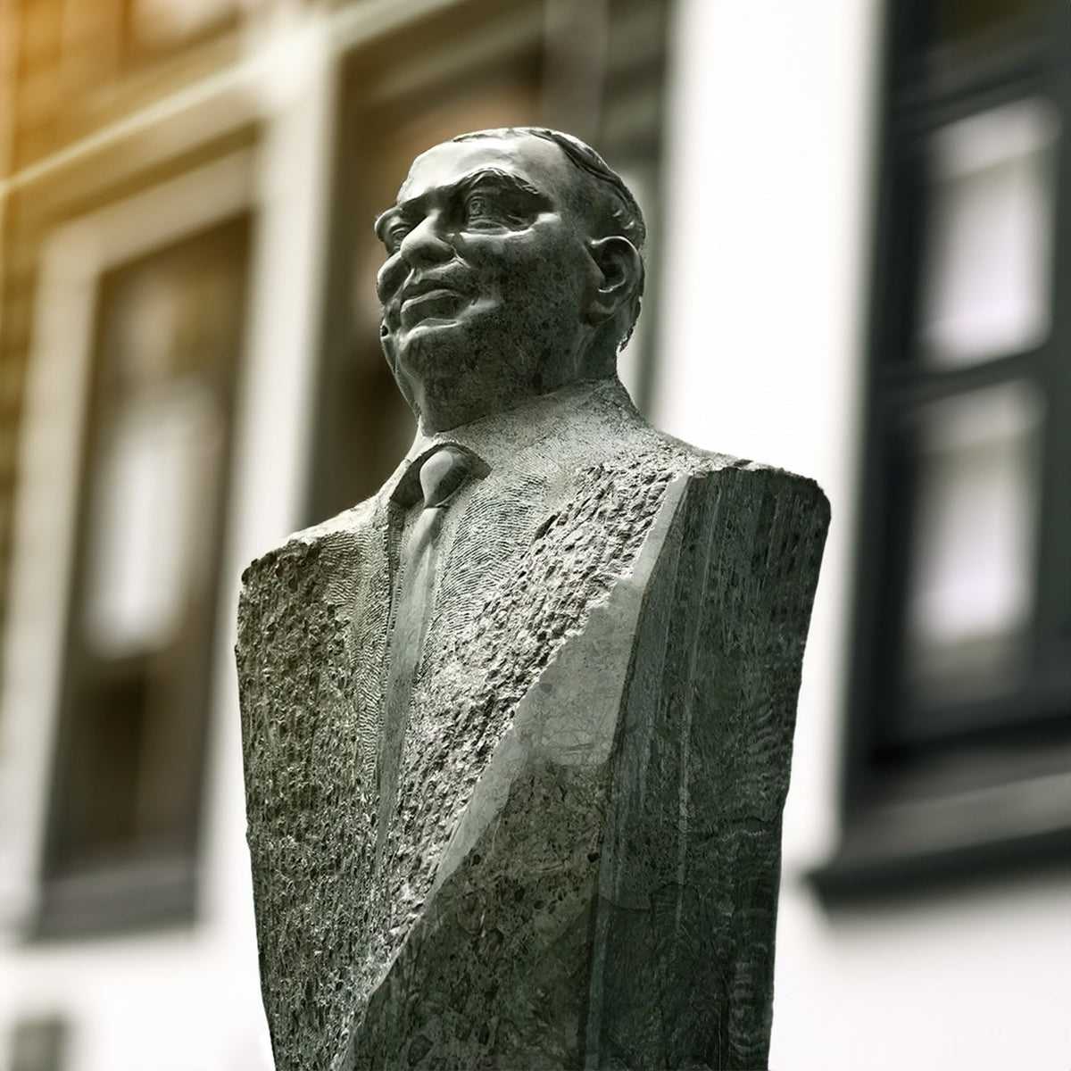 A stone or bronze bust sculpture of a man in a suit, shown outdoors against a softly blurred building facade with windows.