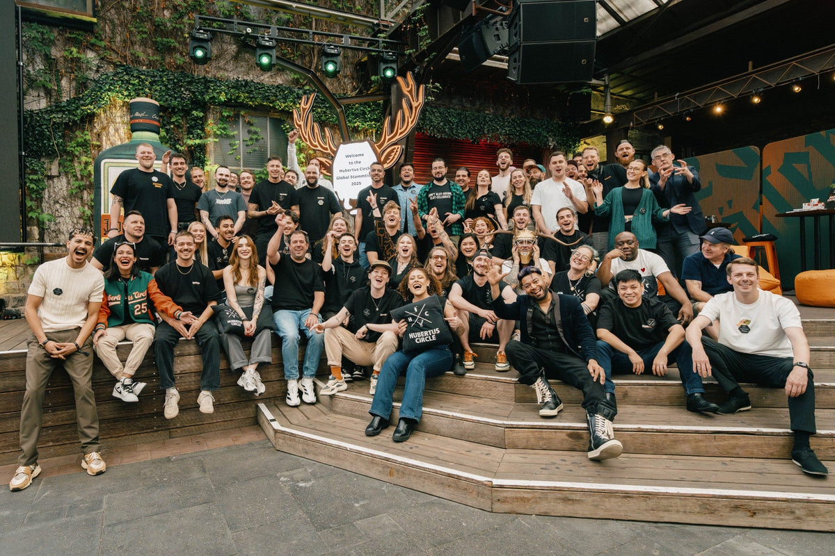 A large group of people posing together on outdoor wooden steps in a rustic, industrial-style venue with stone walls, hanging lights, and greenery.