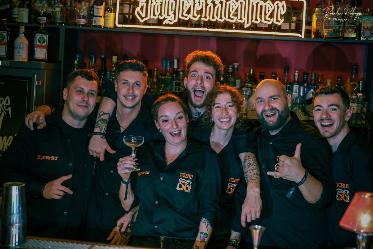 A group of seven cheerful bartenders in black branded "Team 56" shirts pose together behind a bar under a neon Jägermeister sign.