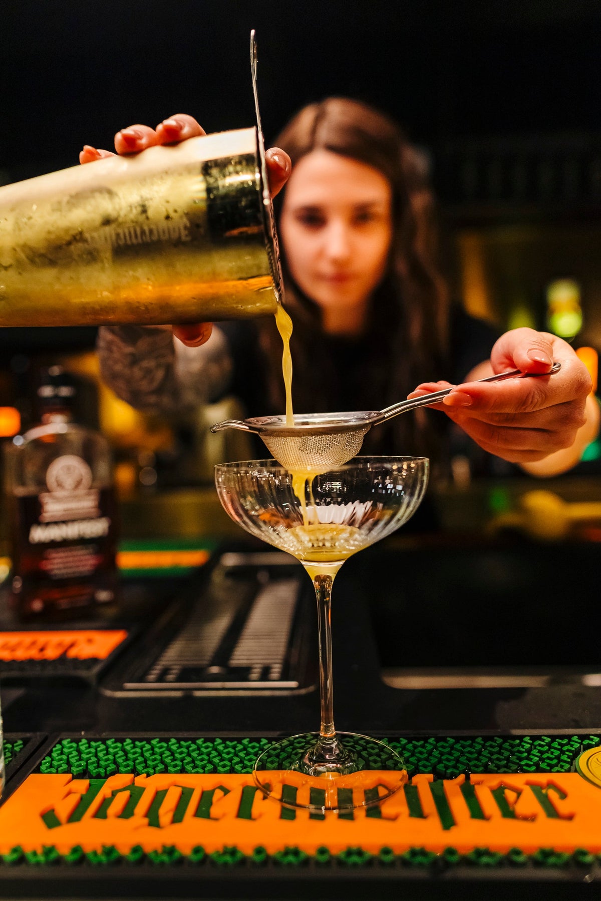 Close-up of a person pouring a yellow cocktail from a metal shaker through a strainer into a stemmed glass on a bar counter, with bottles and bar tools visible in the background.