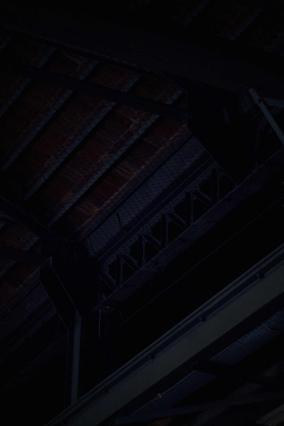 Dark close-up of an industrial ceiling with exposed metal beams, trusses, and brick support structures in very low light.