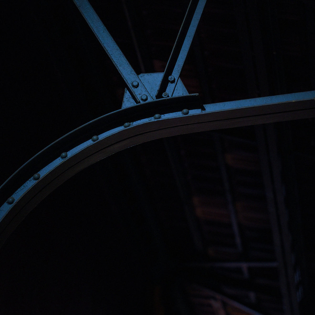 Close-up of a dark metal curved beam or bracket with rivets and bolts against a very dark background.