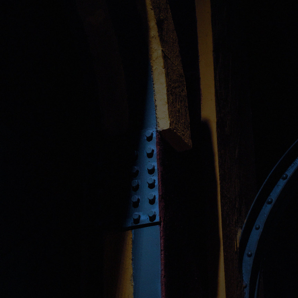 A close-up, low-key photograph of a dark blue metal riveted bracket or support structure against a very dark background with warm yellow-brown vertical accents.