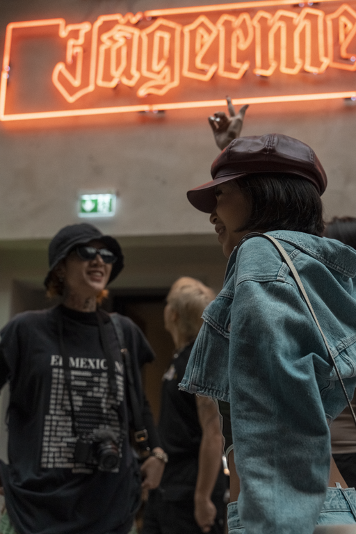 A street-style photograph featuring a person in the foreground wearing a dark brown leather cap and a light blue denim jacket, with another person in a black t-shirt and bucket hat in the background under a glowing orange Jägermeister neon sign in the Jägermeister guesthouse in Wolfenbüttel.