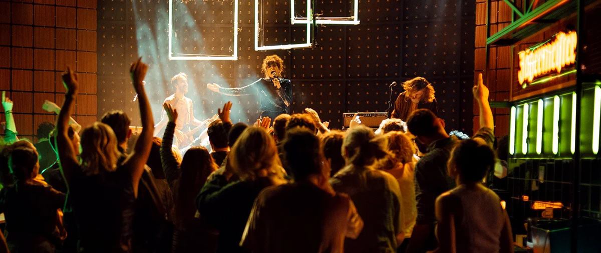 A live concert scene with a performer on stage under dramatic stage lighting, with a crowd of people raising their hands in the foreground.