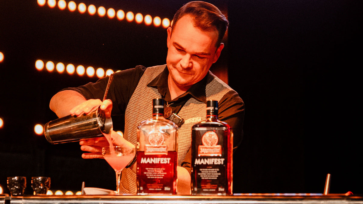 A bartender in a black shirt and patterned vest pours a drink behind two bottles of Jägermeister Manifest liquor on a bar counter, with warm stage lights in the background.