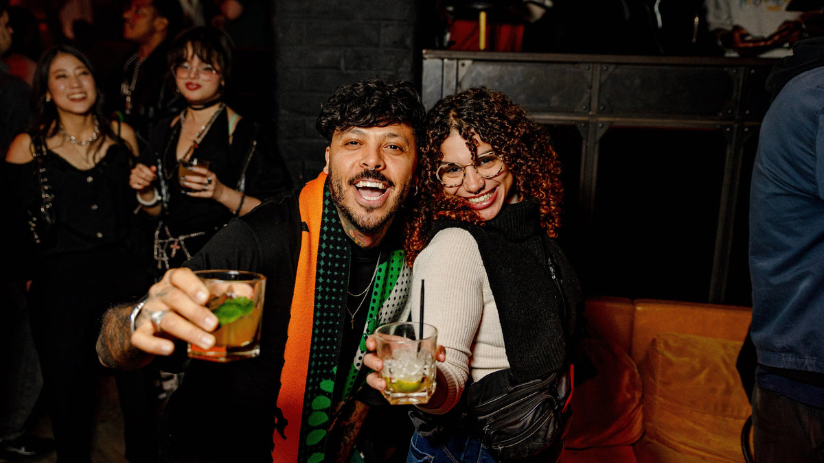 A man and a woman posing together at a social event or party, holding cocktails. The man is wearing a black shirt with a bright orange and green striped scarf, and the woman is wearing a black top.