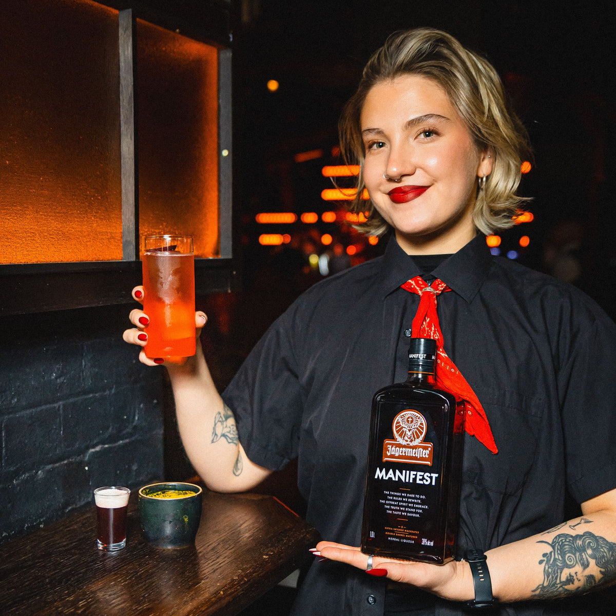 A woman in a black shirt holding a bottle of Jägermeister Manifest and a drink in a dimly lit bar setting.