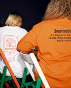 Two people seated on green and white chairs, shown from behind wearing Jägermeisters branded T-shirts. One shirt is white with an orange label print, and the other is bright orange with the Jägermeister HQ address in black.