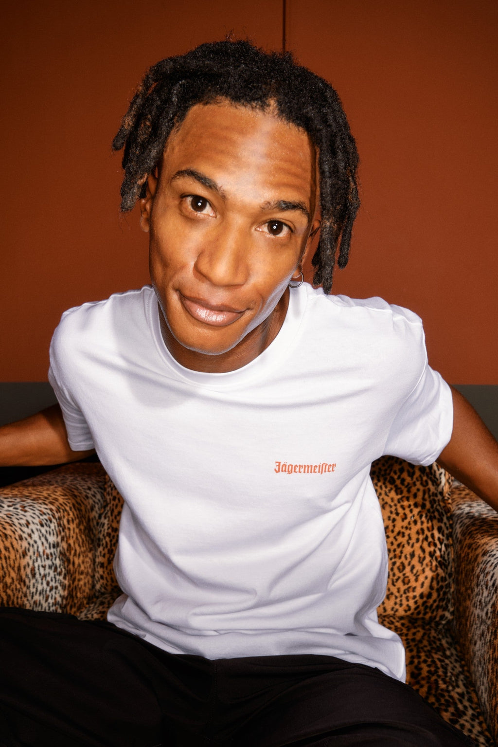 Person wearing a white t-shirt with a Jägermeister logo on the chest, sitting on a patterned chair against an orange wall.