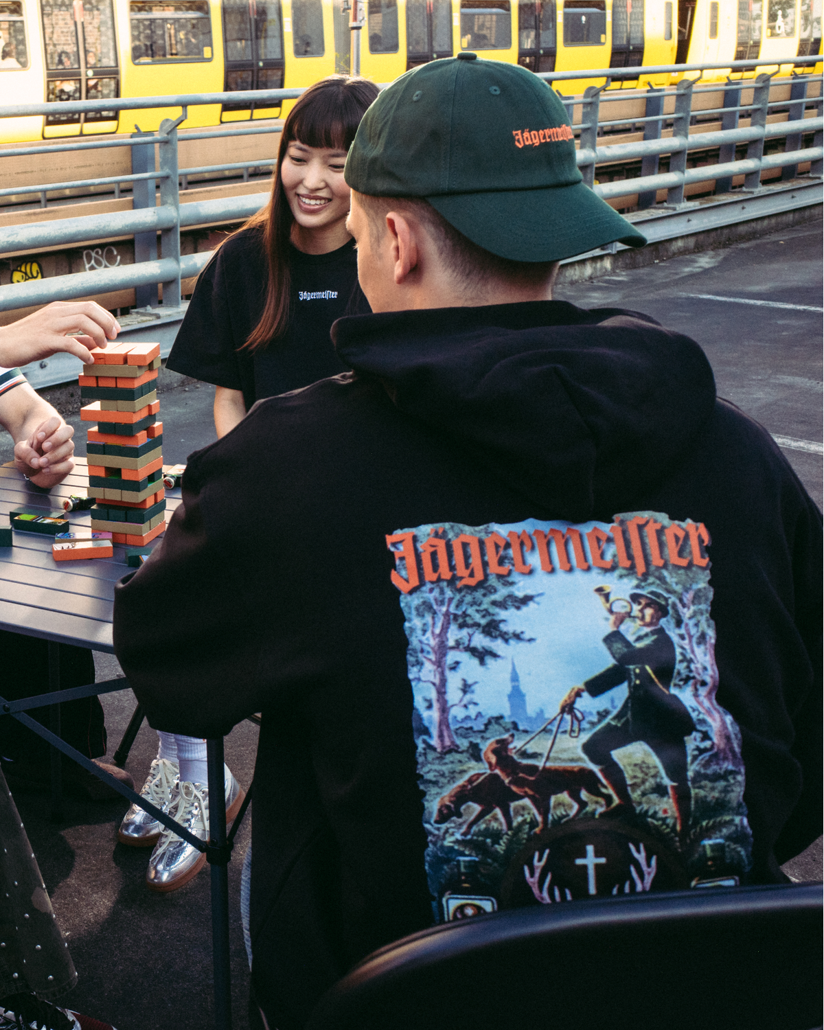 Four people sitting around a folding table outdoors, playing a game of stacking blocks. A yellow and white train is visible in the background behind a metal railing. The person in the foreground is wearing the Jägermeister Tradition Hoodie and a green Jägermeister Dad Cap.