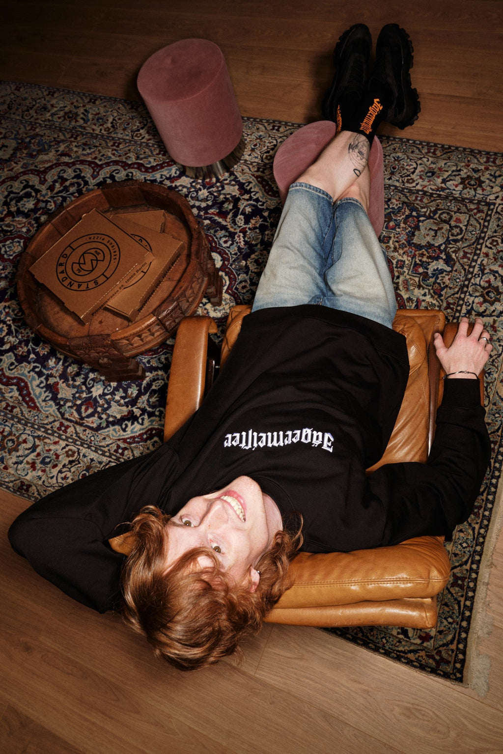 Persoon ligt op een leren stoel met de voeten op een tapijt, en draagt een zwarte Jägermeister Crewneck.