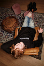Persoon ligt op een leren stoel met de voeten op een tapijt, en draagt een zwarte Jägermeister Crewneck.