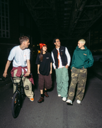 Vier mensen die lopen en fietsen op een donkere straat in de nacht onder een verhoogd treinspoor, gekleed in Jägermeister streetwear in verschillende kleuren.