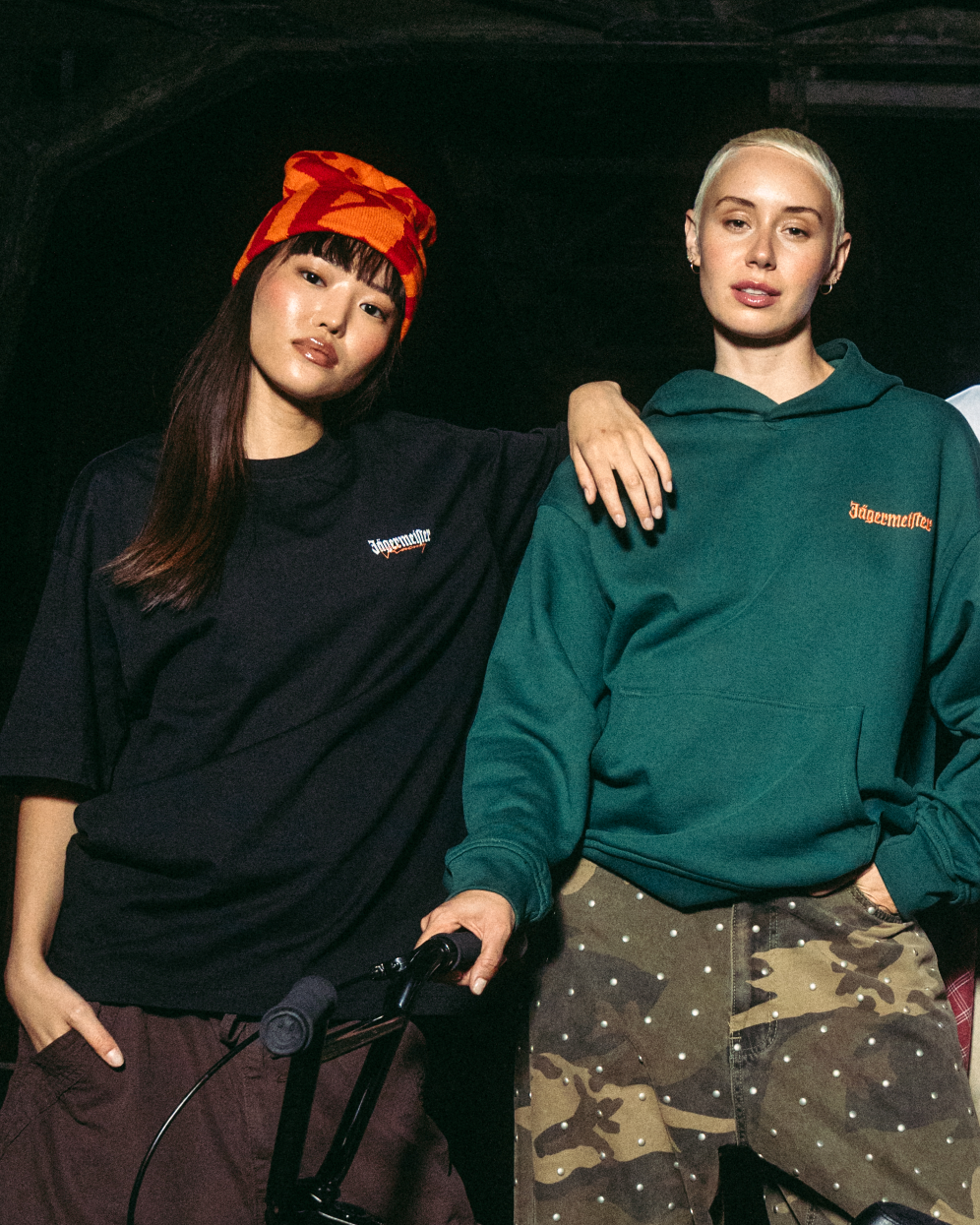 Twee jonge vrouwen poseren met een zwarte BMX-fiets voor een donkere industriële brug bij nacht. Ze dragen Jägermeister streetwear, waaronder een groene hoodie, wit T-shirt, zwart vest en cargobroeken.