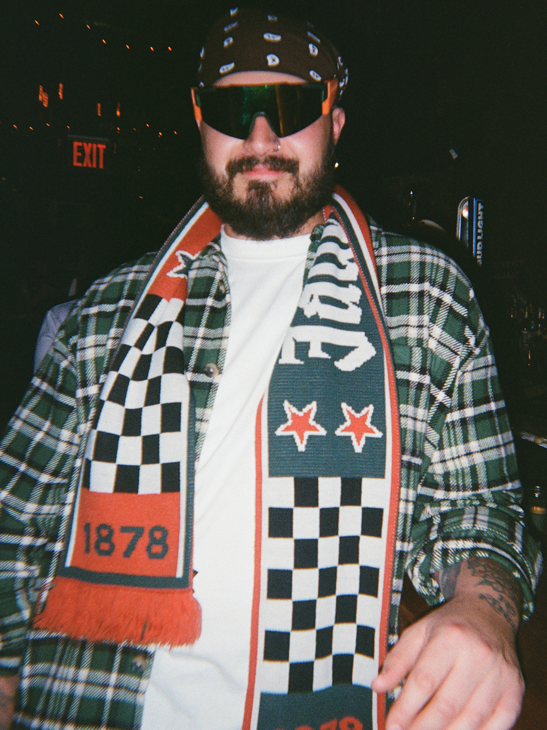Person wearing a plaid jacket with a Jägermeister Racing scarf, sunglasses, and a cap.