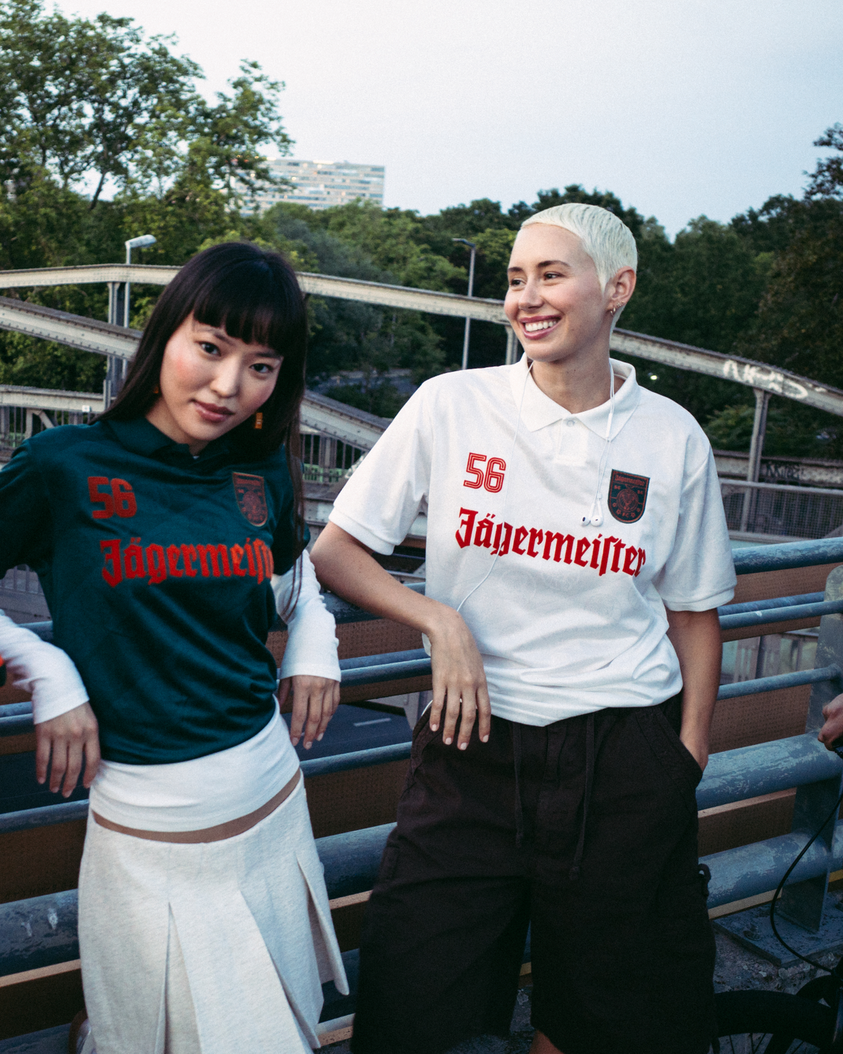 Four people standing outdoors on a bridge or walkway, wearing streetwear. One person wears a bright orange zip-up jacket, another wears a dark green Jägermeister jersey with a white skirt, a third wears a white Jägermeister jersey with dark shorts, and a fourth wears a black hoodie and an orange cap.