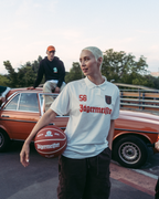 A group of four people posing outdoors on a bridge or overpass with a vintage orange car behind them. The central figure holds a basketball and wears a white Jägermeister jersey, while others wear bright orange, green, and black streetwear.