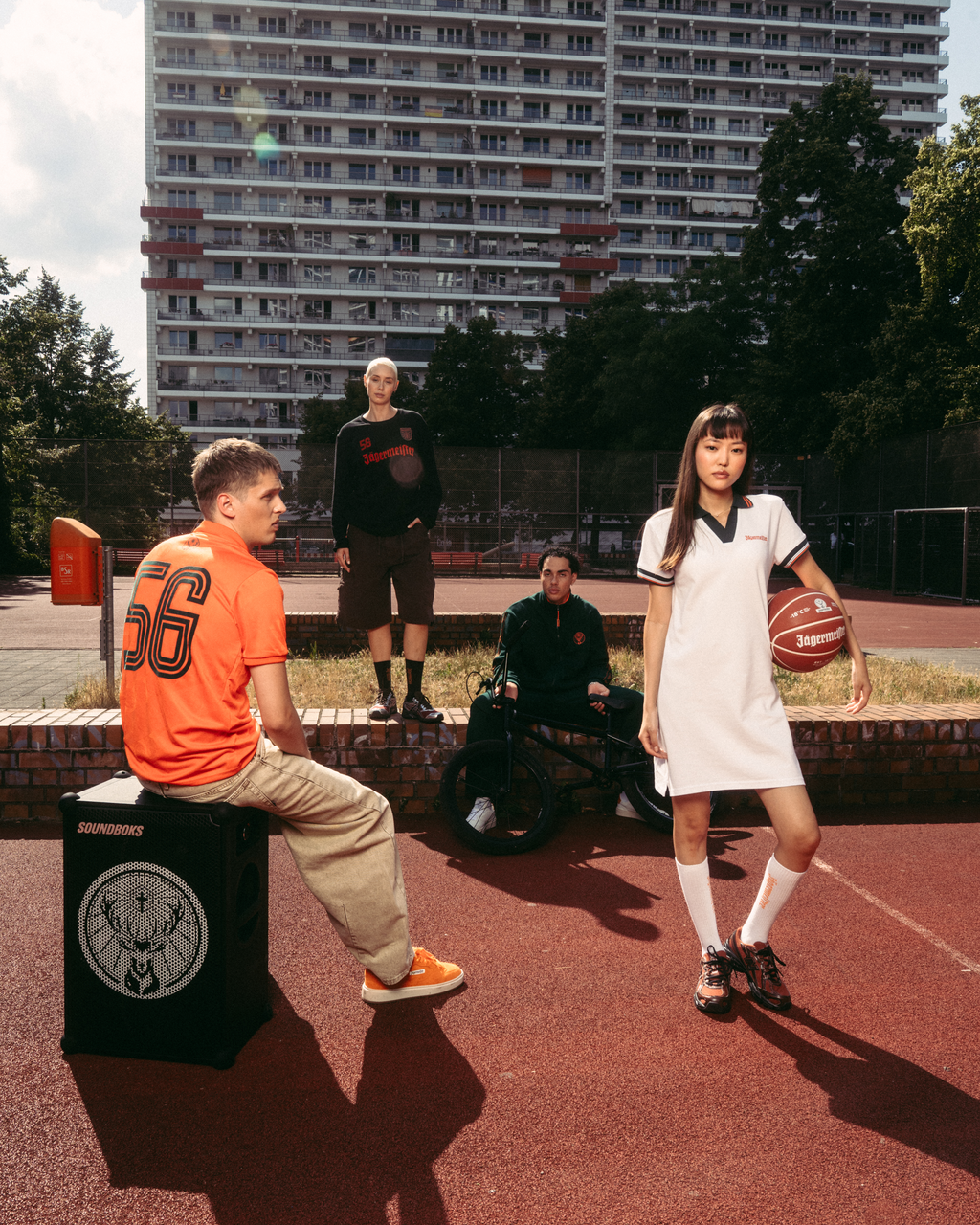 Outdoor streetwear lifestyle scene on a basketball court with a tall apartment building in the background. A seated man in an orange Jägermeister jersey sits on a black speaker-like box, while a standing woman in a white Jägermeister polo dress holds a basketball.