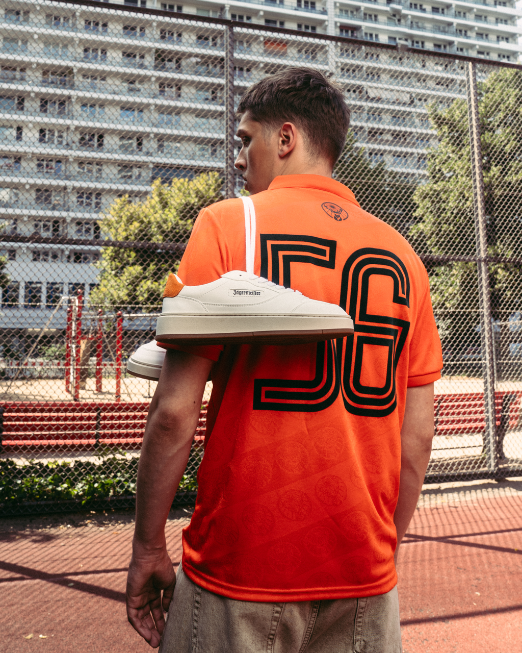 A person stands on an outdoor sports court wearing a bright orange Jägermeister jersey with a large “56” printed on the back. A pair of white Jägermeister sneakers is draped over one shoulder, with a chain-link fence and apartment buildings in the background.