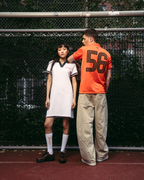 Two models standing on a red tennis court in front of a chain-link fence. The model on the left wears a white athletic Jägermeister polo dress with dark green trim, white socks, and black shoes.