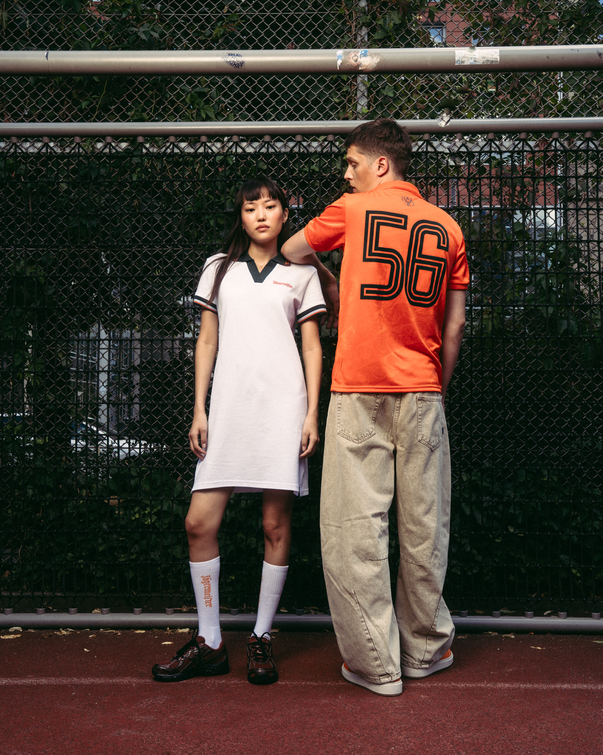 Two models standing on a red tennis court in front of a chain-link fence. The model on the left wears a white athletic Jägermeister polo dress with dark green trim, white socks, and black shoes.