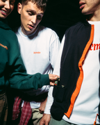 Three young men standing close together in a dark setting, wearing Jägermeister apparel. One wears a dark green hoodie, another wears a white long-sleeve tee, and the third wears a black and orange zip-up jacket over a white tee.