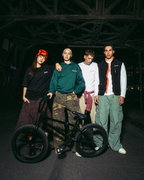 Four young men posing with a black BMX bike in a dark, industrial bridge setting at night. They are dressed in Jägermeister streetwear outfits, including a green hoodie, white t-shirt, black vest, and cargo pants.
