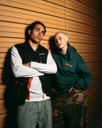 Two models posing against a warm wood-paneled wall. The model on the left wears a black Jägermeister vest over a white long-sleeve shirt with orange accents, and olive cargo pants.