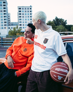 Two people standing beside a vintage red car in an outdoor parking area at dusk. One man wears a bright orange  Jägermeister sweatshirt and black pants, while the woman wears a white Jägermeister jersey and dark pants and holds a basketball.
