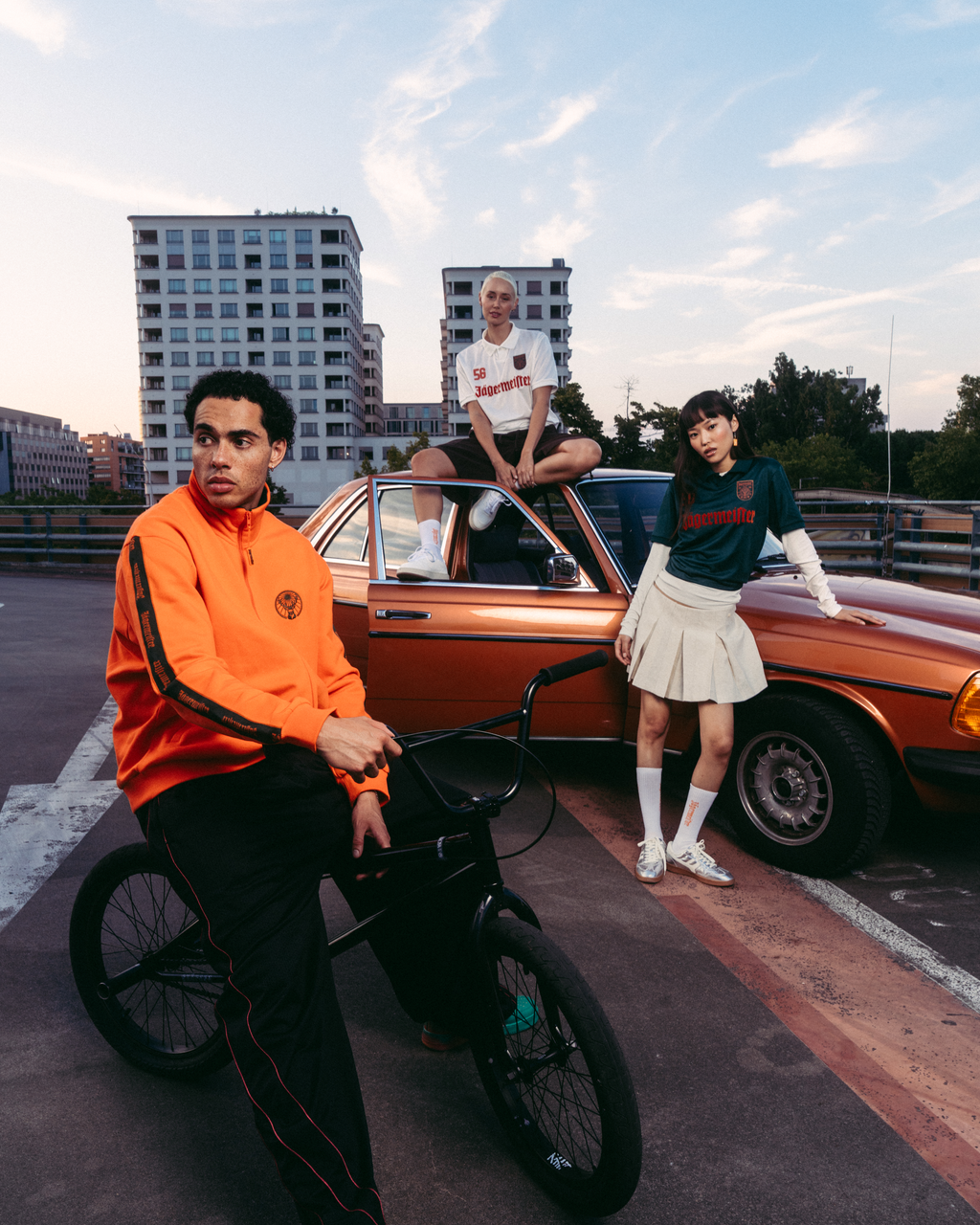 Three models in Jägermeister streetwear outfits posing with a vintage orange car and a black BMX bike on an urban rooftop at sunset. The outfits include a bright orange Jägermeister zipper jacket, black track pants with red stripes, a white Jägermeister jersey, a green Jägermeister jersey with orange branding and a white pleated skirt.