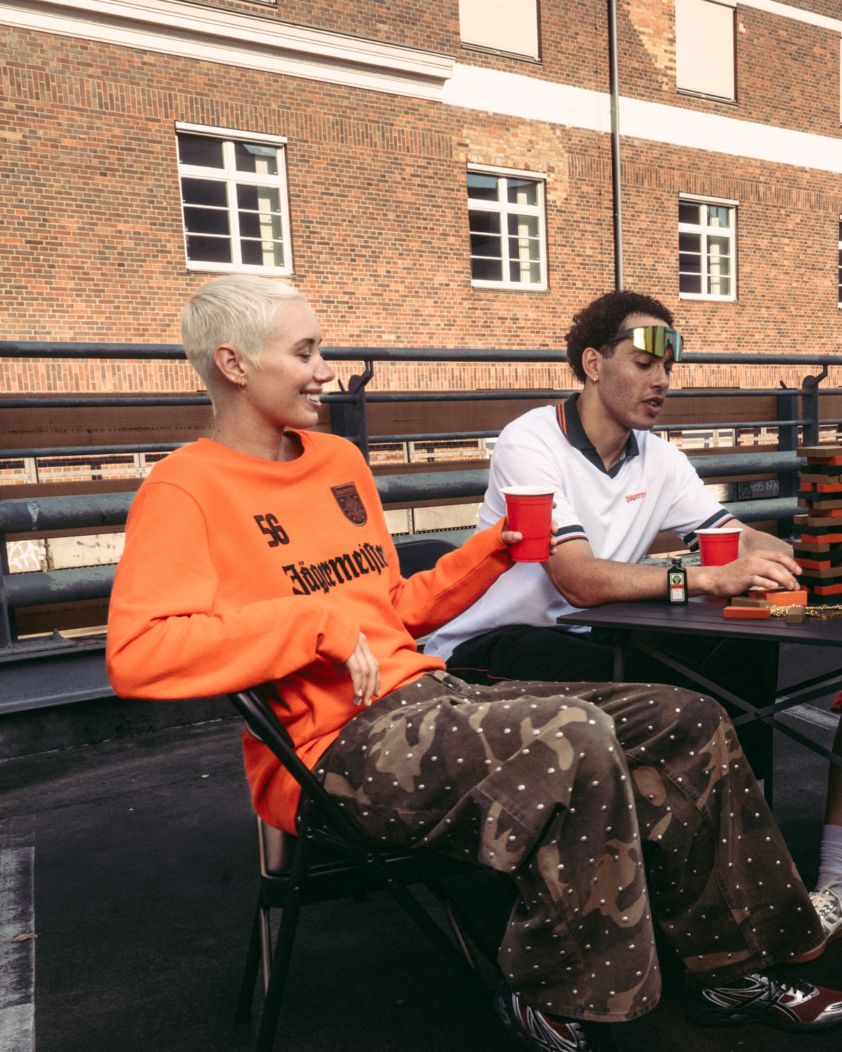Four people sitting around a small table on an outdoor rooftop or bridge, holding red cups. They are dressed in casual Jägermeister streetwear, including an orange longsleeve and a white polo shirt.