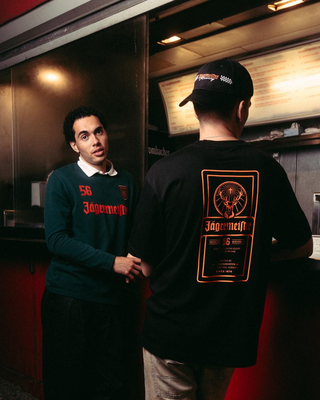 Two men standing outside a red food truck at night. One man faces the camera wearing a dark green Jägermeister longsleeve with orange text, black pants, and white sneakers.