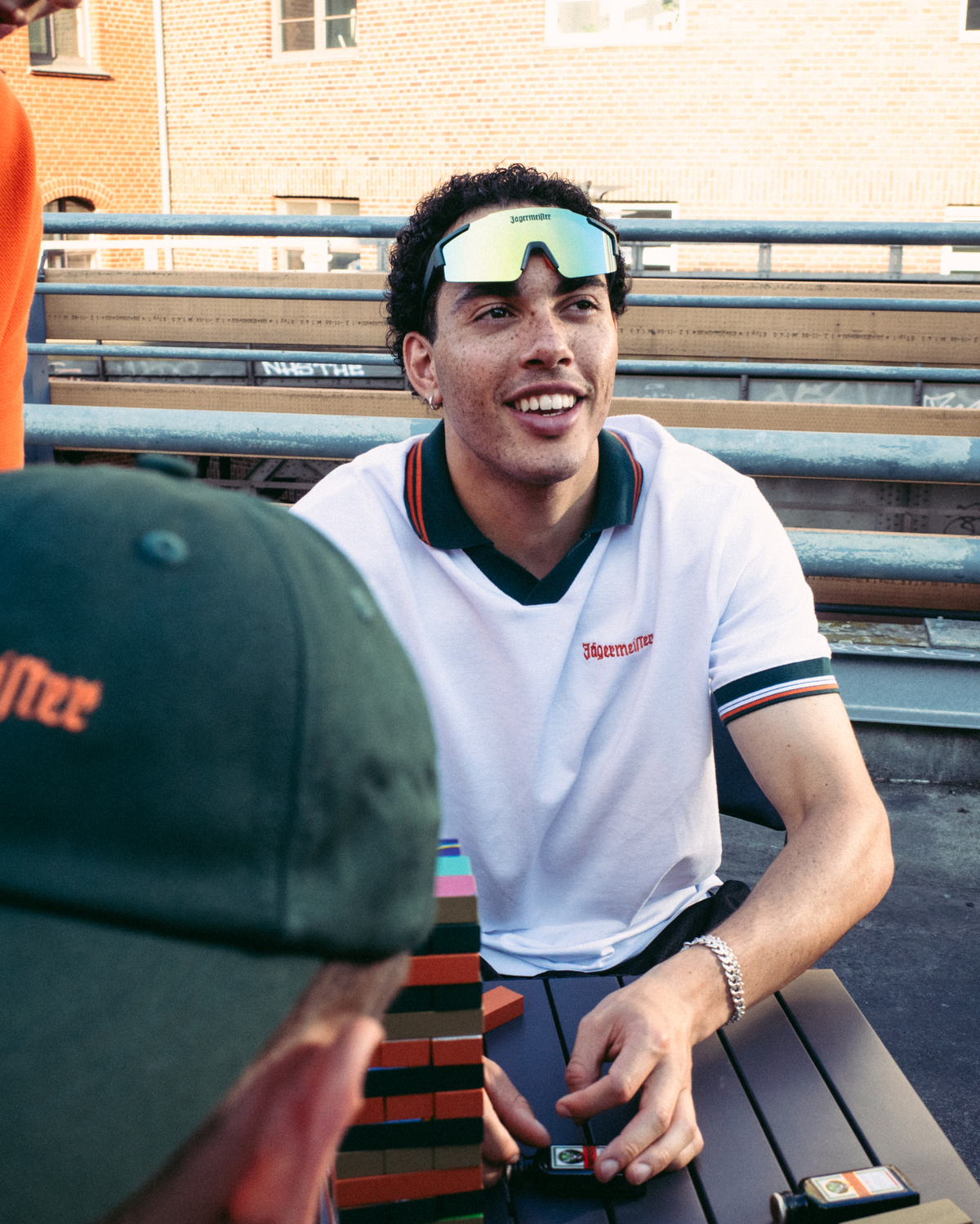 A man in a white and green Jägermeister polo shirt smiles while sitting outdoors at a table, with another person in a green Jägermeier cap in the foreground and a brick building in the background.