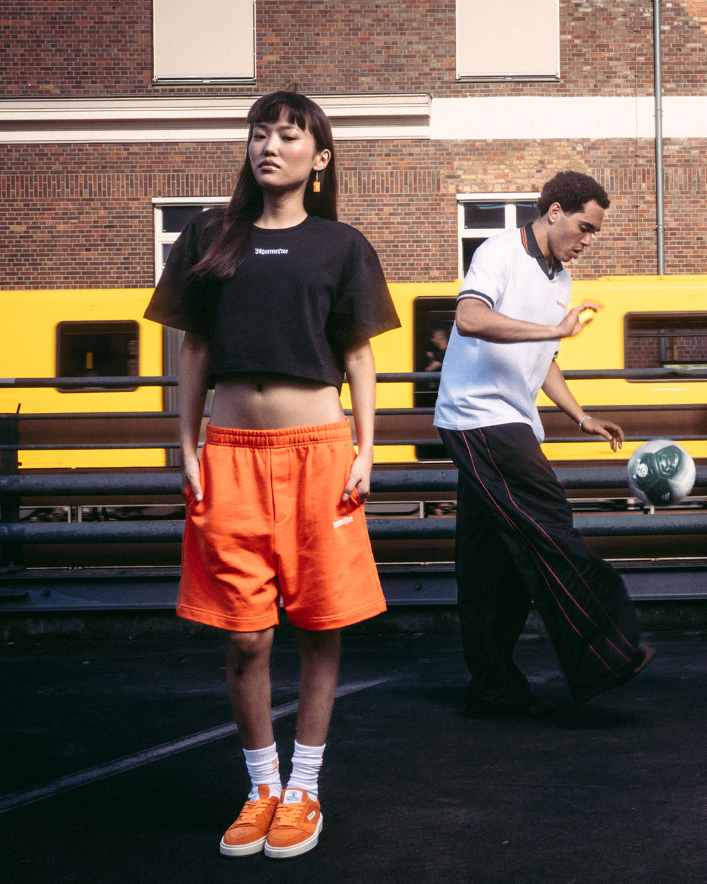Vier modellen in Jägermeister streetwear, waaronder oranje shorts, een zwarte crop top, een wit T-shirt en een zwarte hoodie met een grafische print op de rug, staand op een donkere verharde ondergrond voor een bakstenen gebouw en een gele trein.