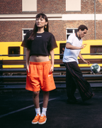 Vier modellen in Jägermeister streetwear, waaronder oranje shorts, een zwarte crop top, een wit T-shirt en een zwarte hoodie met een grafische print op de rug, staand op een donkere verharde ondergrond voor een bakstenen gebouw en een gele trein.
