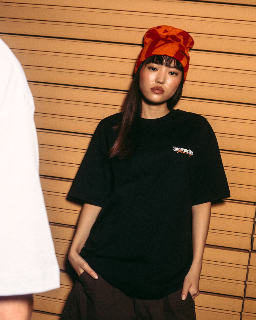 Twee modellen poseren tegen een wand van houten latten. Het model links laat de achterkant zien van een wit T-shirt met een grote oranje opdruk met daarop 'Jägermeister'.