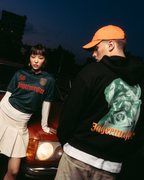Two models posing in front of a vintage red car at dusk. One model wears a dark green 'Jägermeister' jersey with a white pleated skirt, and the other wears a black Jägermeister Waldi hoodie and an orange cap.