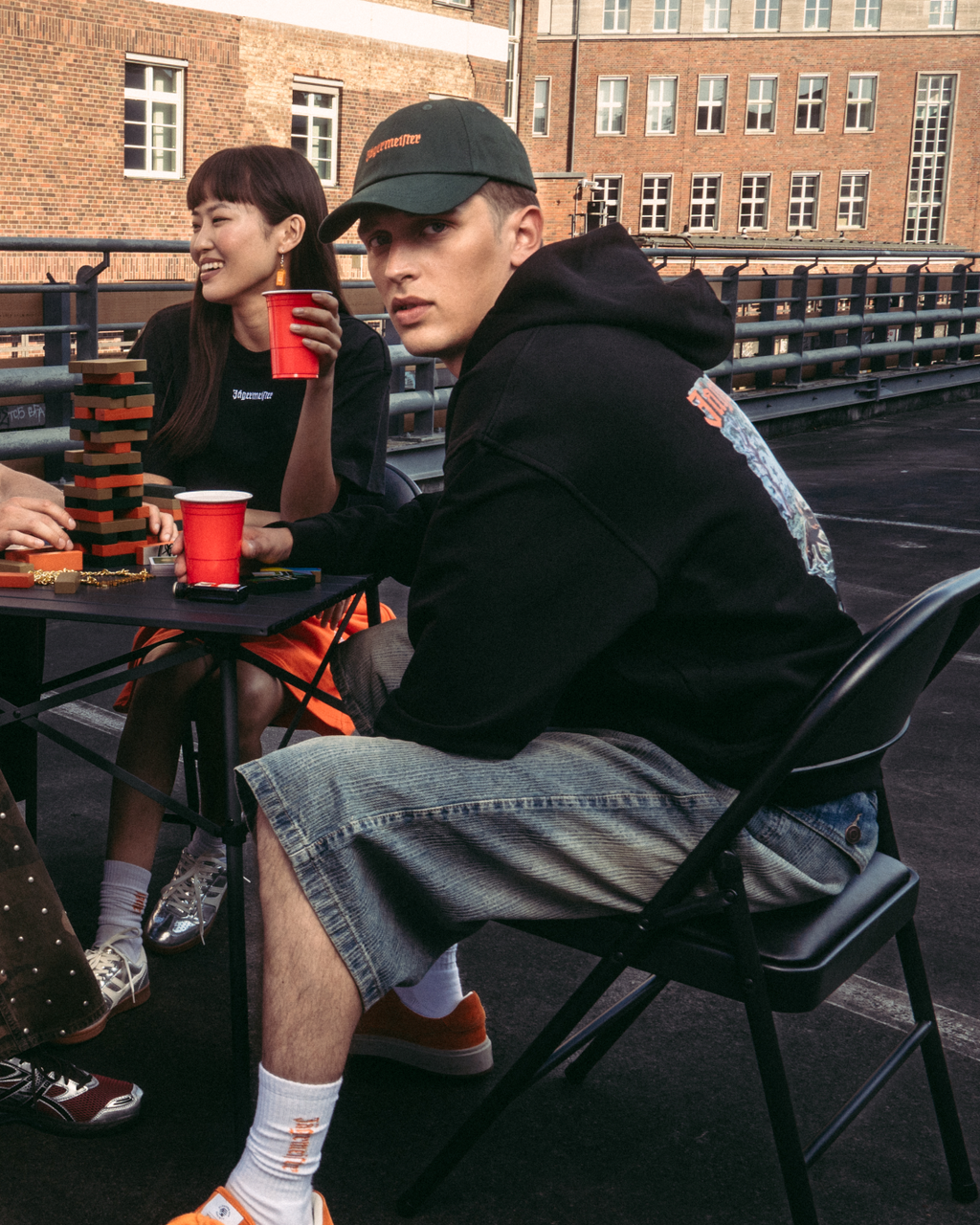 Four people sitting around a small table on an outdoor rooftop or bridge, holding red cups. They are dressed in casual Jägermeister streetwear, including an orange sweatshirt, white t-shirt, black hoodie, and patterned pants.