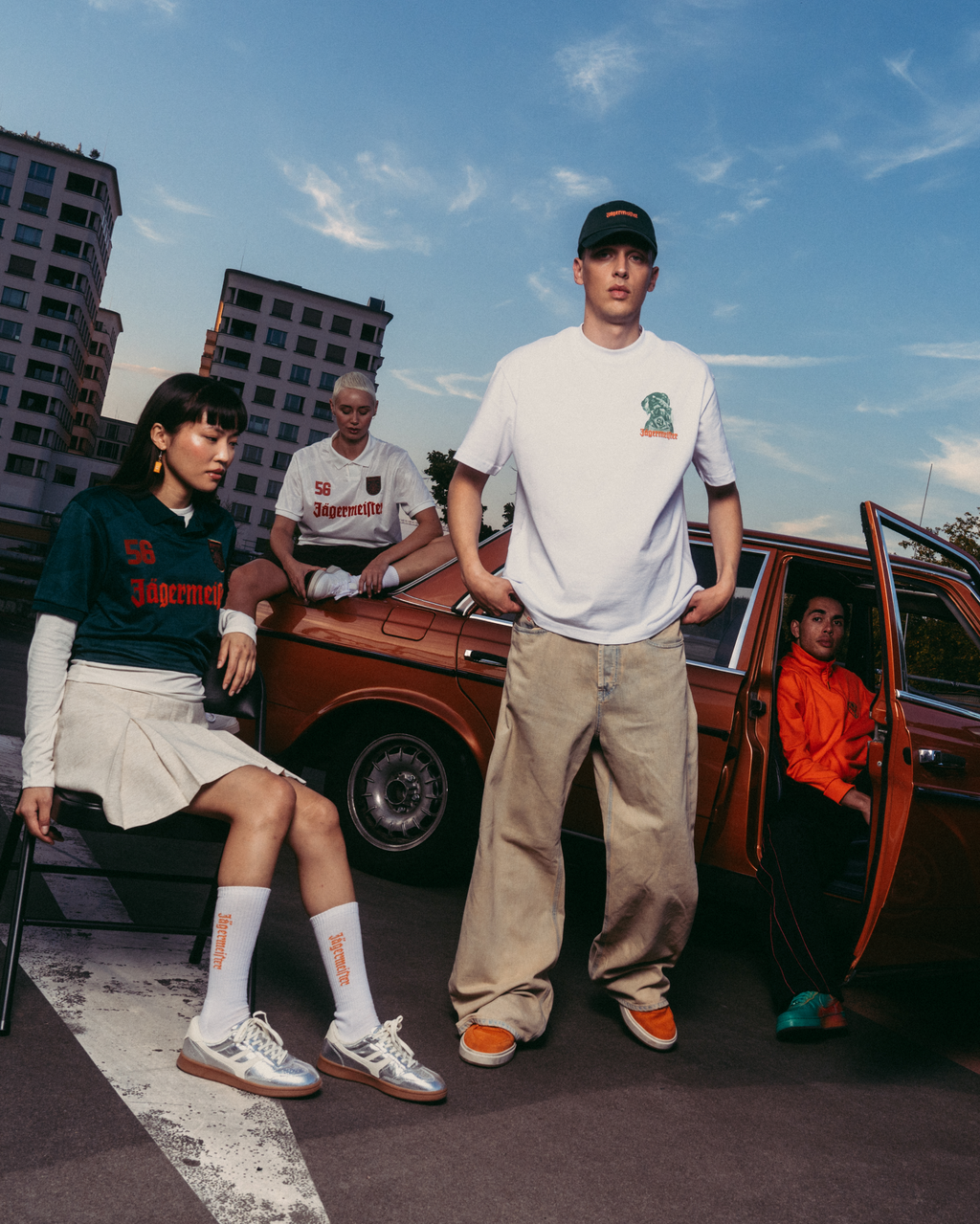 Drie modellen poseren met een vintage oranjebruine auto op een stadsstraat onder een blauwe lucht. Eén model zit links in een donkergroene sweatshirt en witte rok, één staat in het midden in een wit T-shirt en beige broek, en één zit in de open autodeur met een oranje jas aan.