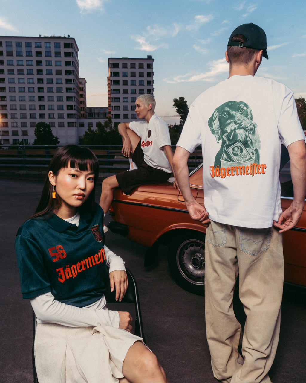 Drie modellen dragen Jägermeister-kleding in een stedelijke buitenomgeving met een vintage oranje auto en appartementen op de achtergrond. Eén model zit op de voorgrond in een donkergroen Jägermeister-shirt, een ander staat met zijn rug naar de camera in een wit T-shirt met graphic en beige broek, en een derde model zit op de auto in een wit T-shirt.