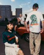 Drie modellen dragen Jägermeister-kleding in een stedelijke buitenomgeving met een vintage oranje auto en appartementen op de achtergrond. Eén model zit op de voorgrond in een donkergroen Jägermeister-shirt, een ander staat met zijn rug naar de camera in een wit T-shirt met graphic en beige broek, en een derde model zit op de auto in een wit T-shirt.