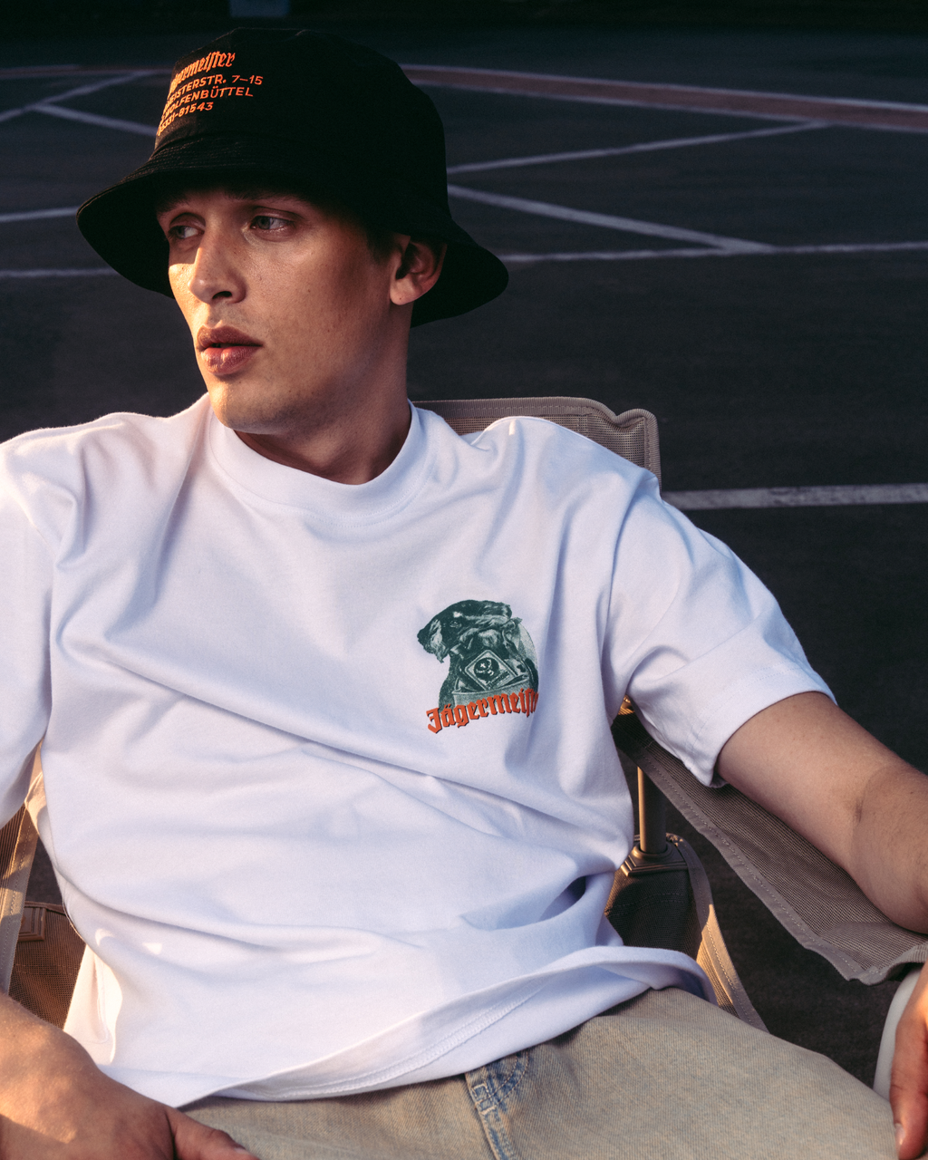 Een man in een wit grafisch T-shirt met een Jägermeister Waldi-logo, een zwarte bucket hat en beige jeans, zittend op een stoel in een buitenomgeving.