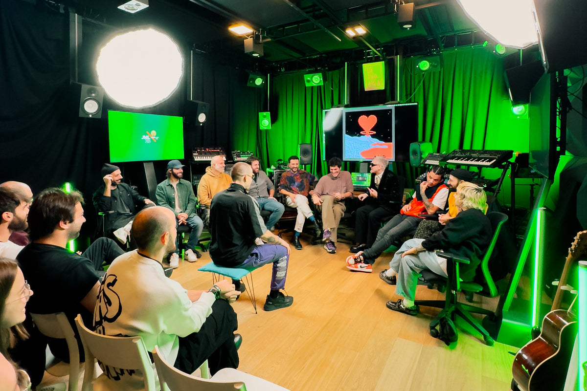 A group of people seated in chairs inside a studio or room with green lighting, screens, microphones, and musical equipment. The room has black curtains, a wood floor, and bright green accent lights.