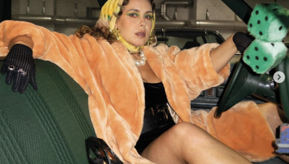 A woman reclining in a vintage car wearing a plush orange coat, black gloves, a yellow headscarf, and a black bodysuit, holding a green dice prop.