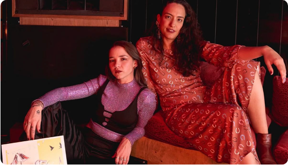 Two women posing together in a dimly lit, warm-toned interior. The woman on the left wears a black top with a sparkling lavender-purple long-sleeve overlay.